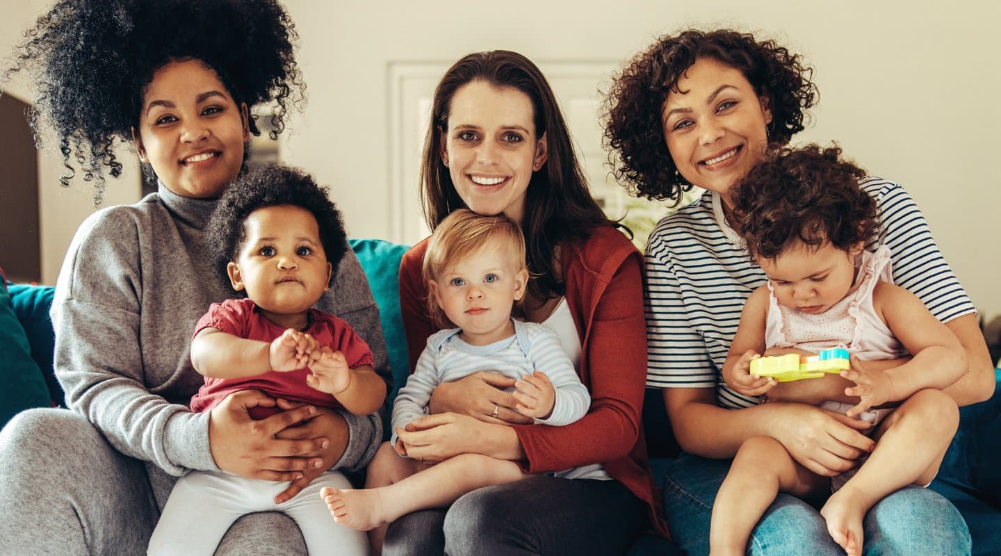 3 women sitting on couch holding babies and smiling