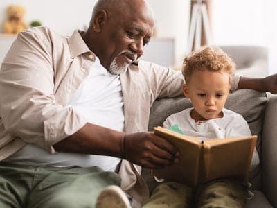 A grandfather reads a book with his grandchild on a couch