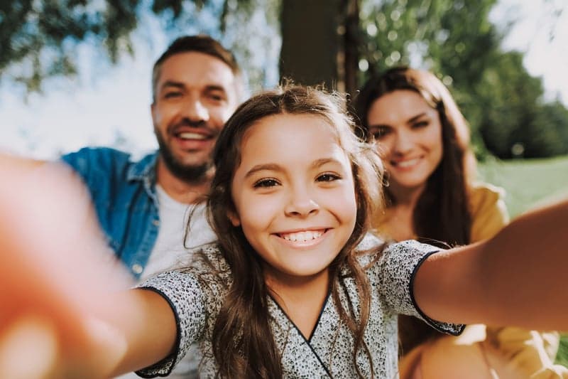 Waist up of smiling girl is making photo with her young parents in green park