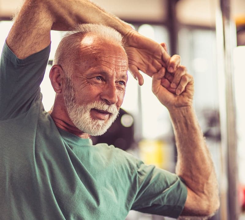 Older man with a white beard stretches his hand over his head and feels joint pain