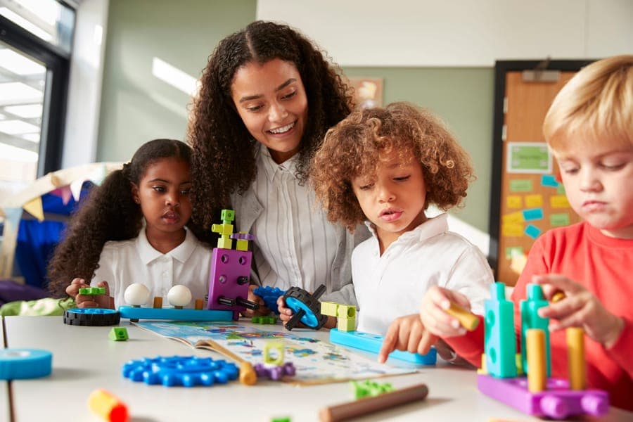 Female teacher sitting at table in play room with three kindergartne children constructing, selective focus