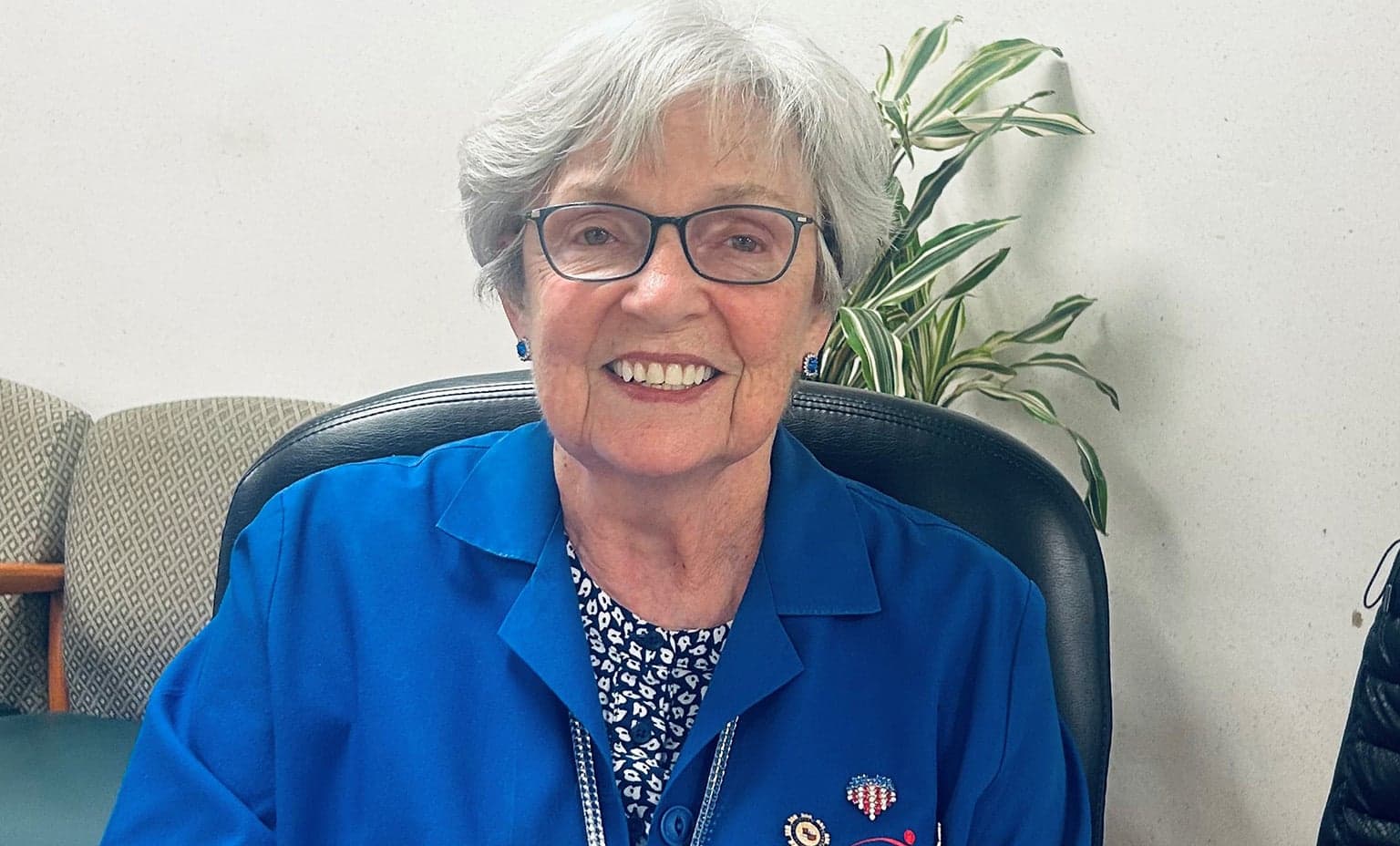 Helen Tselepis wears her blue volunteer coat and smiles while sitting at a desk