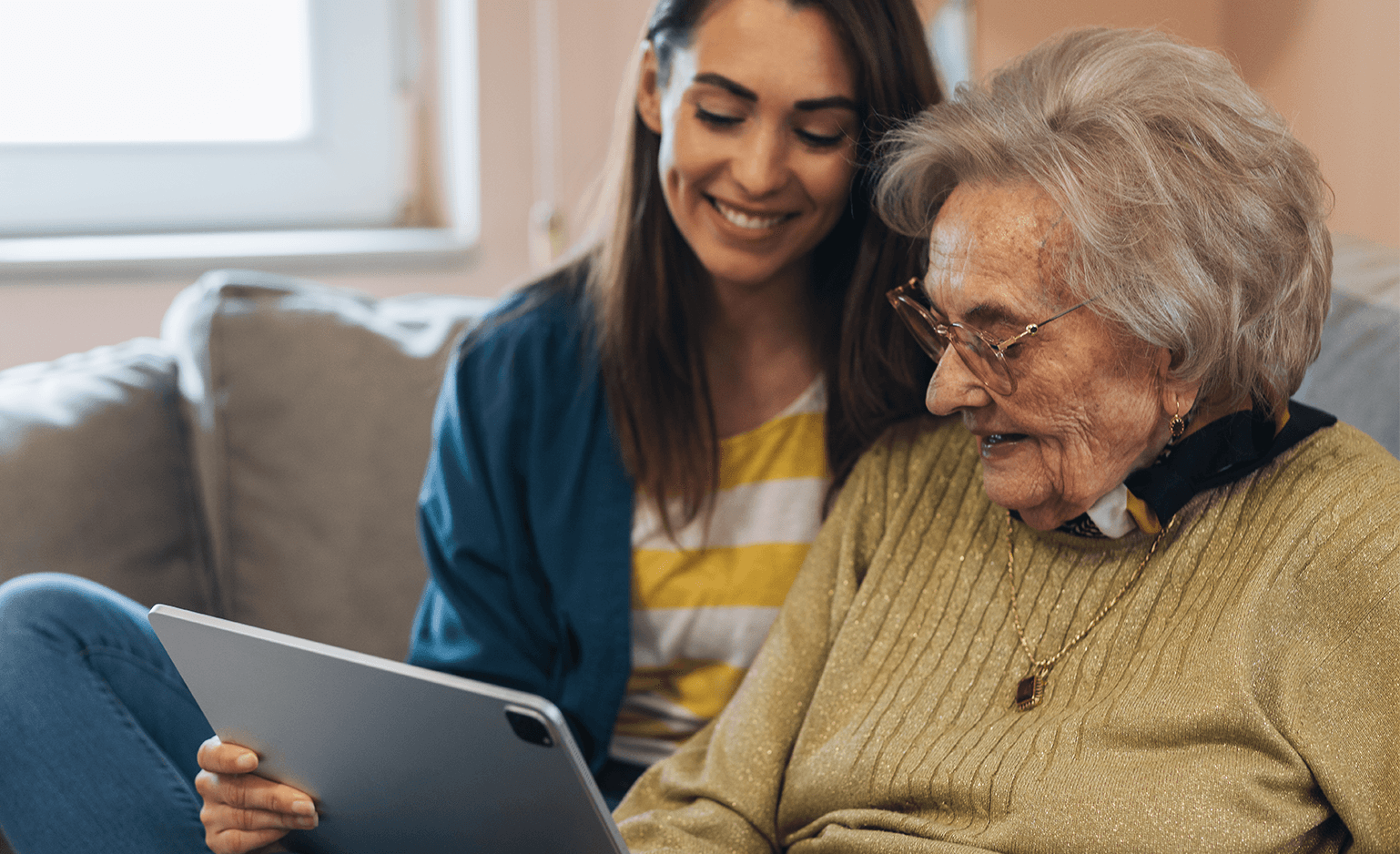 woman and senior woman sitting on couch holding and looking at a tablet.