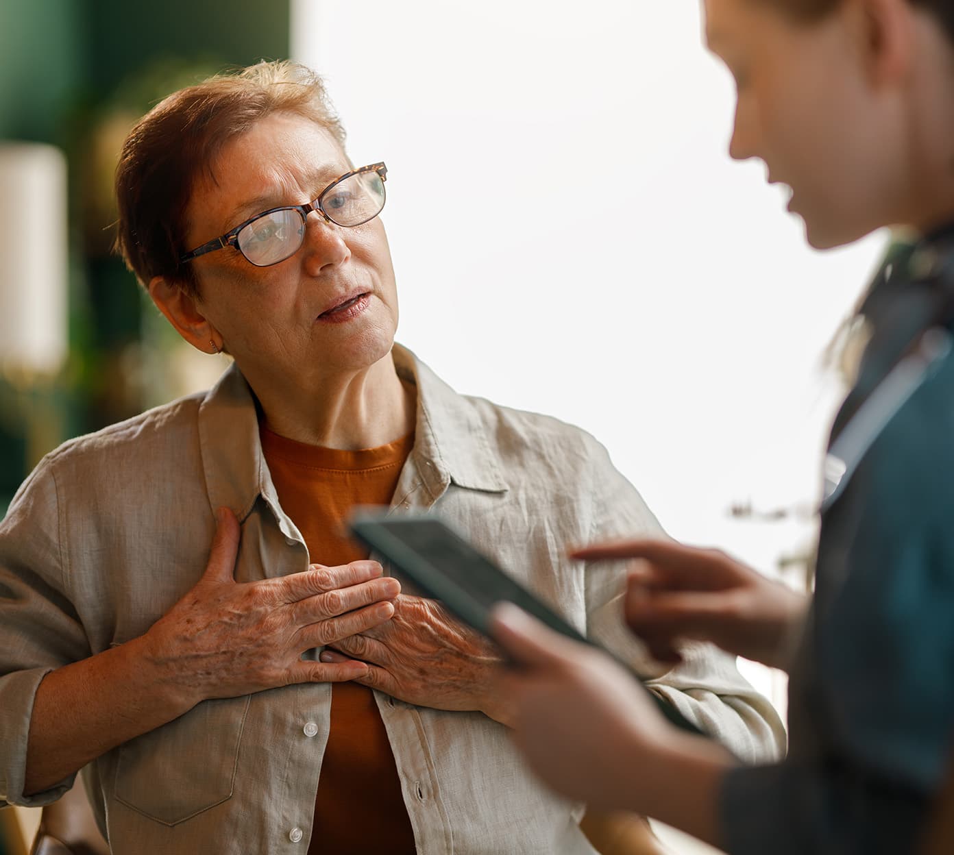 Woman with glasses touches her chest while listening to a someone reading from a clipboard