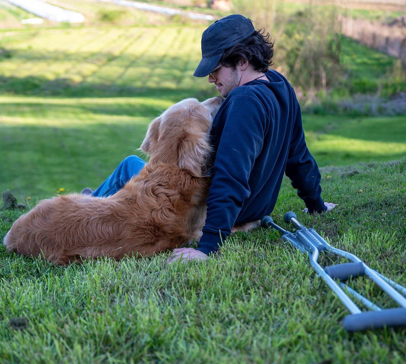 Man sits on a hill with a golden retriever and a pair of crutches is next to them