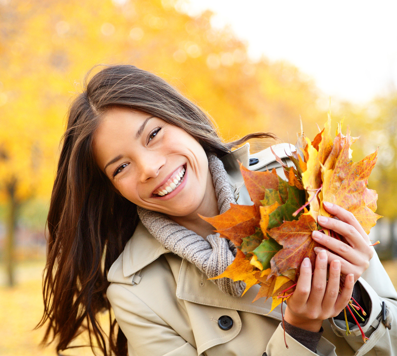 A woman with brown hair wearing a trench coat holds fall leaves