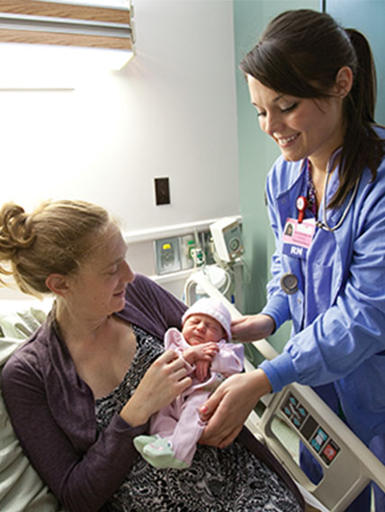 New mother in hospital bed holds infant with nurse standing next to the bed