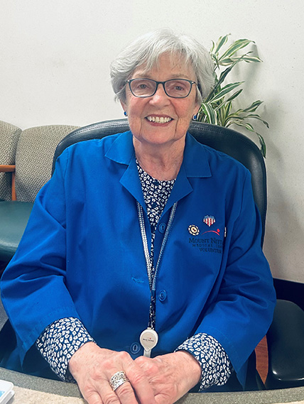 Helen Tselepis wears her blue volunteer coat and smiles while sitting at a desk