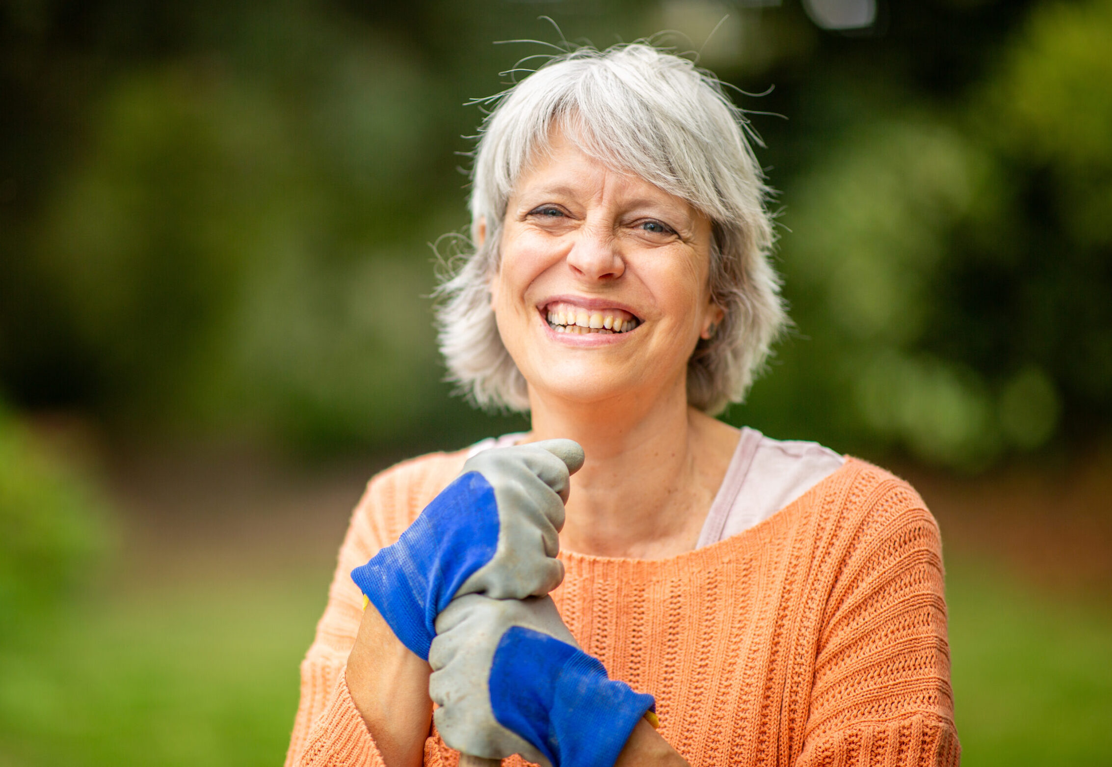 Senior woman with short gray hair, wearing an orange sweater and blue gardening gloves, smiling while standing in a garden