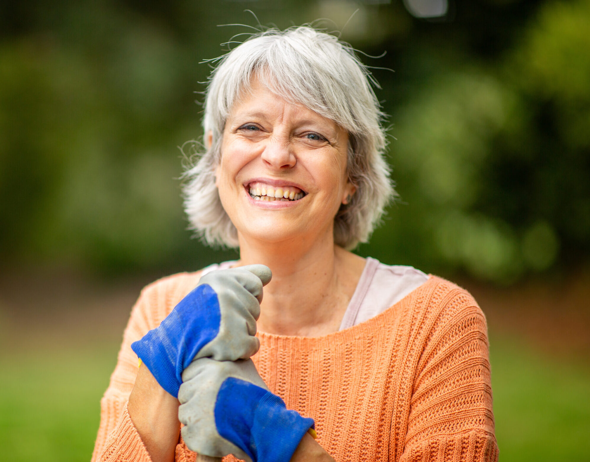 Senior woman with short gray hair, wearing an orange sweater and blue gardening gloves, smiling while standing in a garden