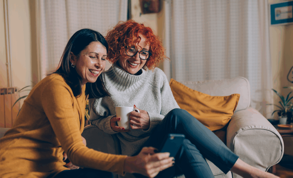 two middle aged women sitting on couch in living room. One woman showing other woman something on her phone, both woman smiling.