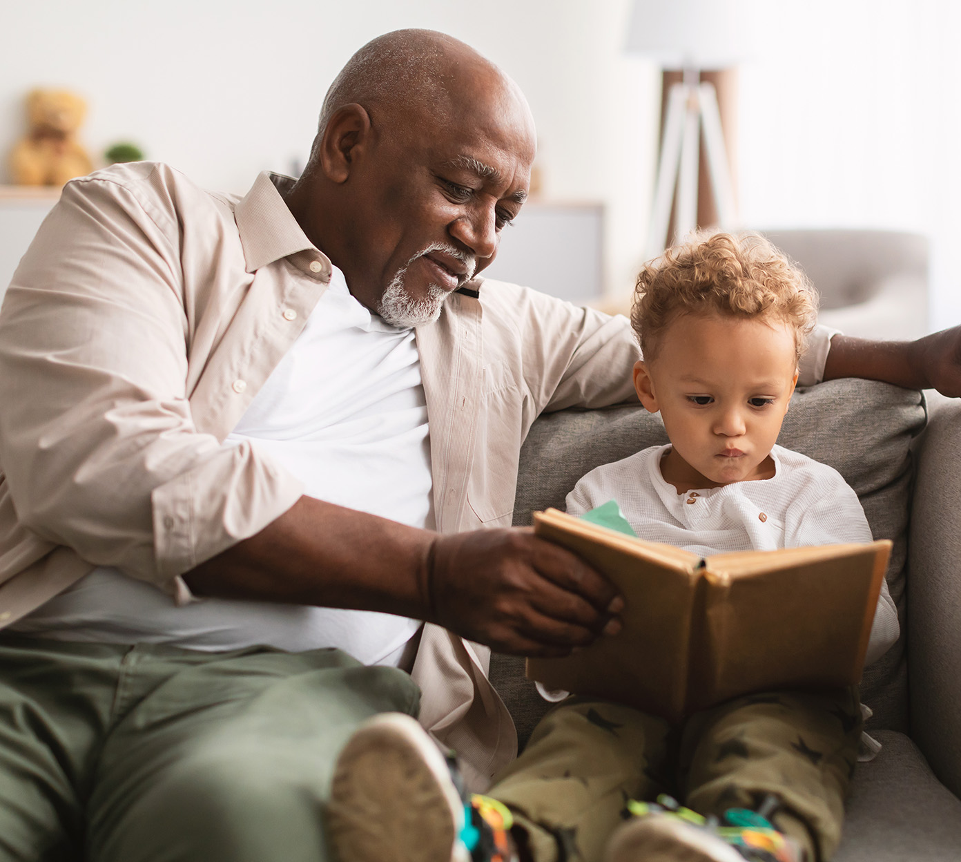 A grandfather reads a book with his grandchild on a couch