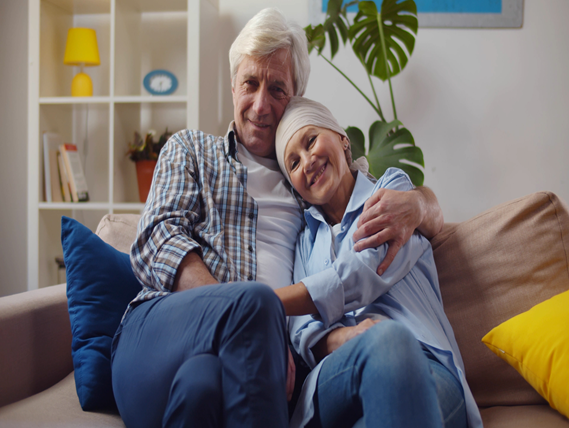 Smiling woman with cancer hugging husband sitting on couch at home.