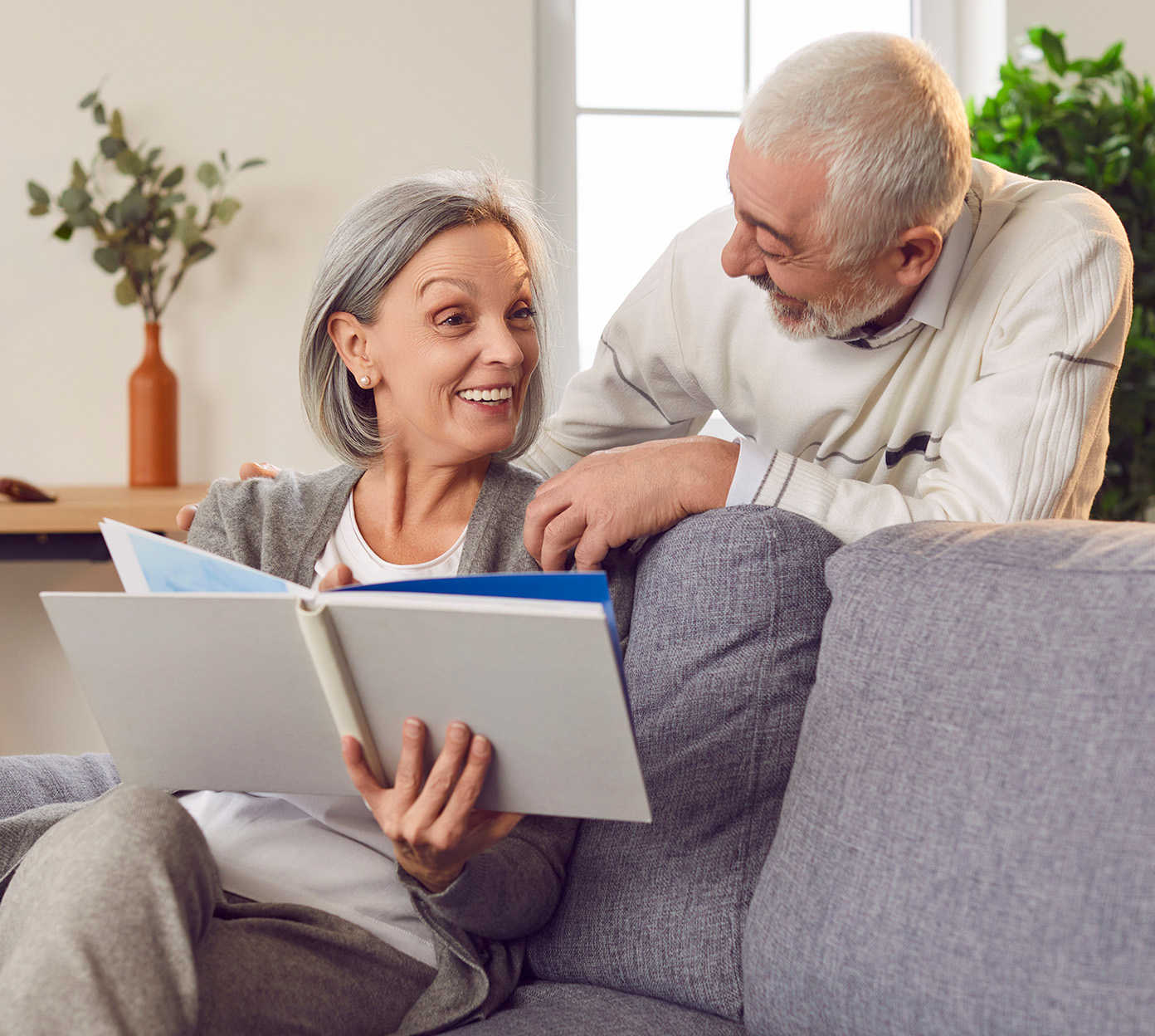 An older woman sits on a couch showing a book to a mature man standing behind her