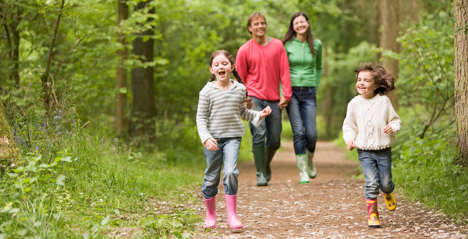 Two kids wearing rain boots on a hiking path with their parents behind them